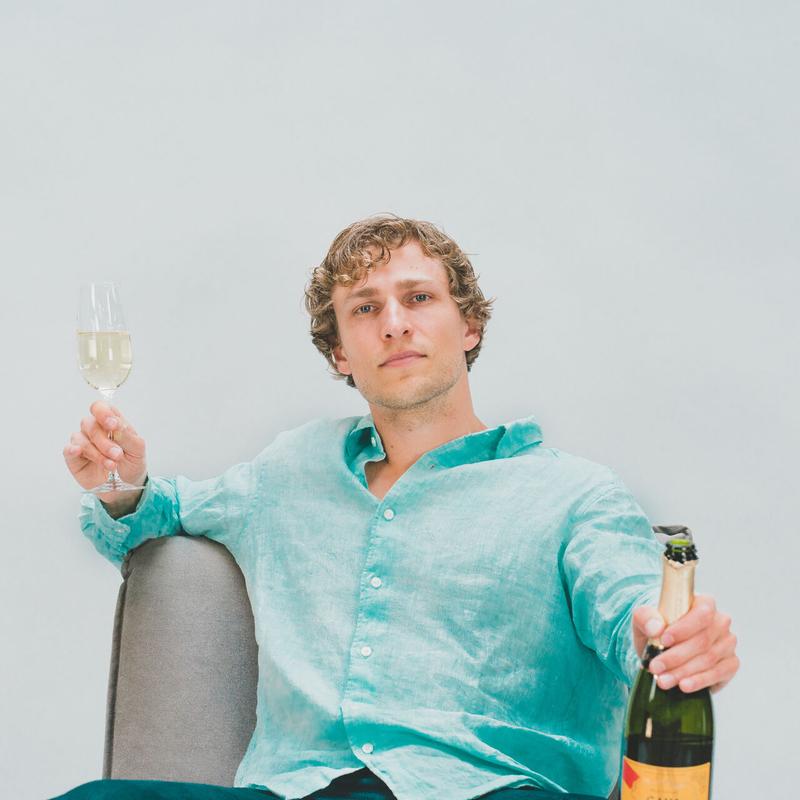Champagne cover art: Dylan Hand holding a glass of campagne in his right hand and a bottle of champagne in his left hand wearing a light green button-up linen shirt in a chair in front of a white background