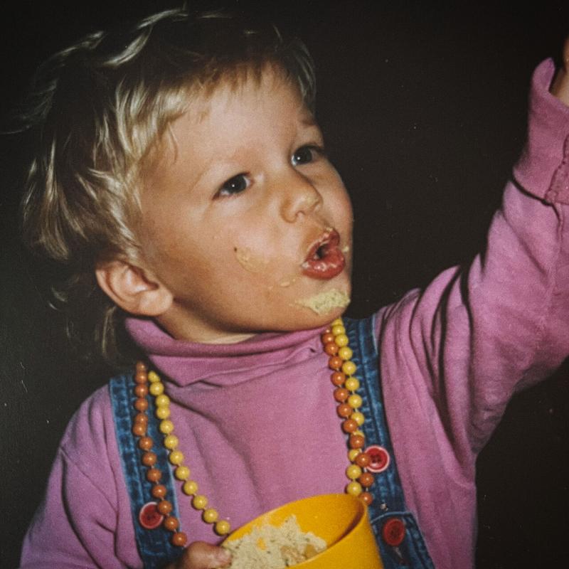 Fresh Eyes cover art: Dylan Hand as a toddler looking up and to the right with his hand outstretched, wearing a pink turtleneck, overalls, orange and yellow beads, and plenty of food on his face.