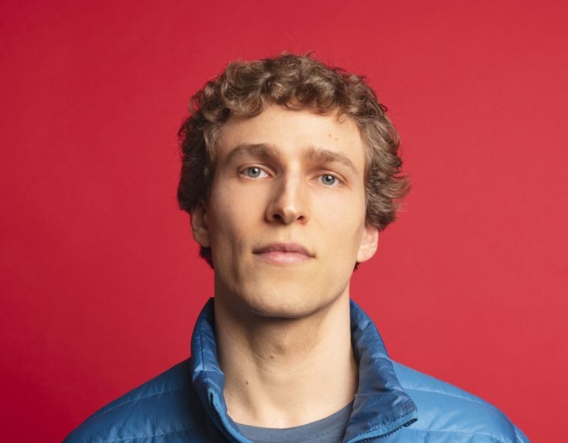 Dylan Hand in front of a bright red background wearing a blue jacket