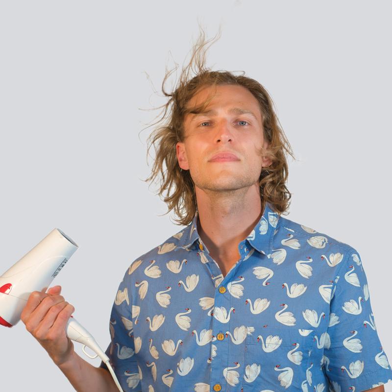 Automatic cover art: Dylan Hand in front of a white background wearing a blue button up shirt covered in swans, holding a hair dryer in his right hand which is blowing his hair up.