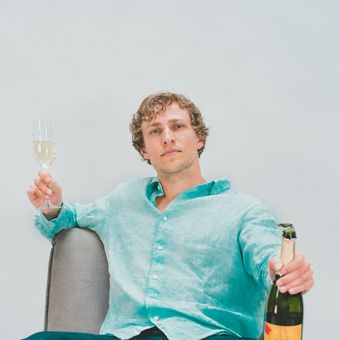 Champagne cover art: Dylan Hand holding a glass of champagne in his right hand and a bottle of champagne in his left hand wearing a light green button-up linen shirt in a chair in front of a white background